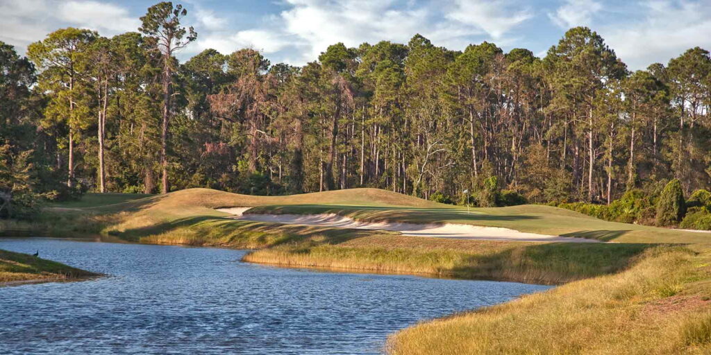 a golf course near a body of water with brown trees under a cloudy sky